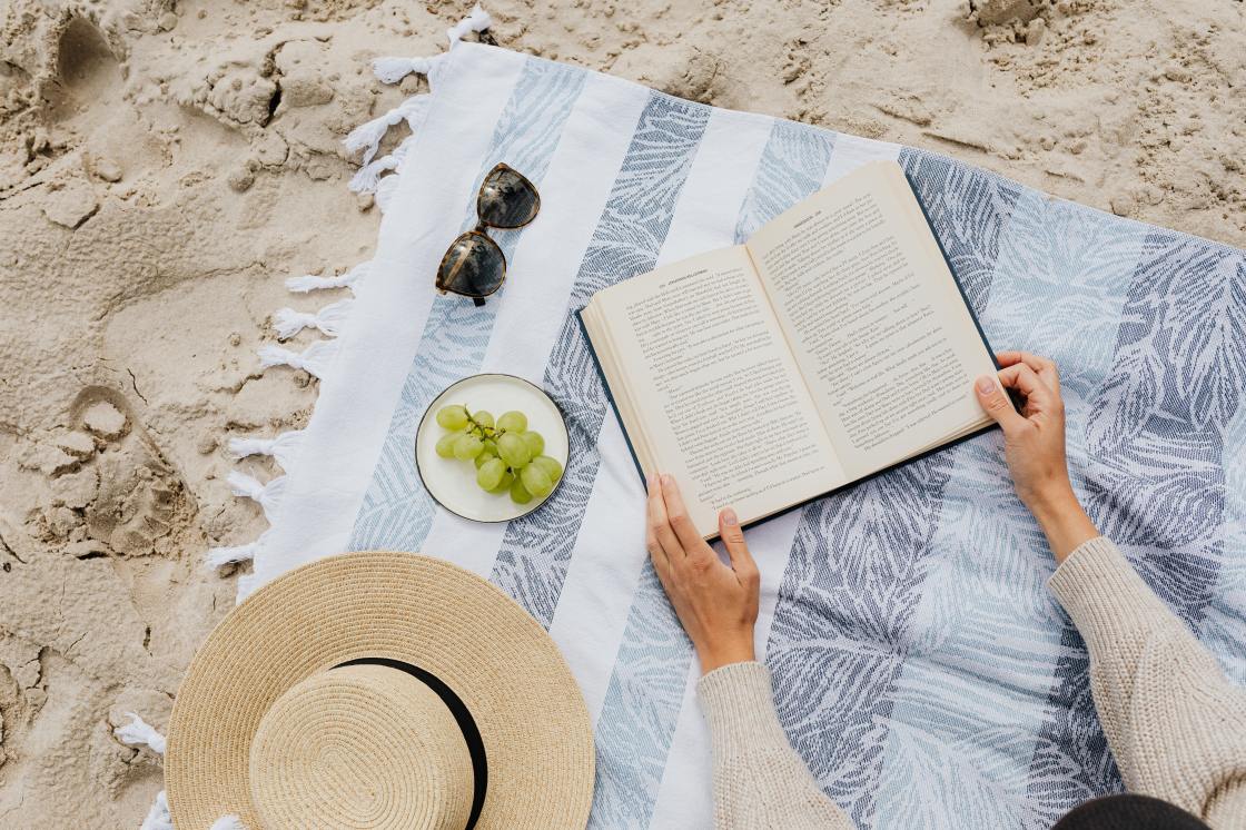 Person reads book by the beach on a blue and white striped towel.