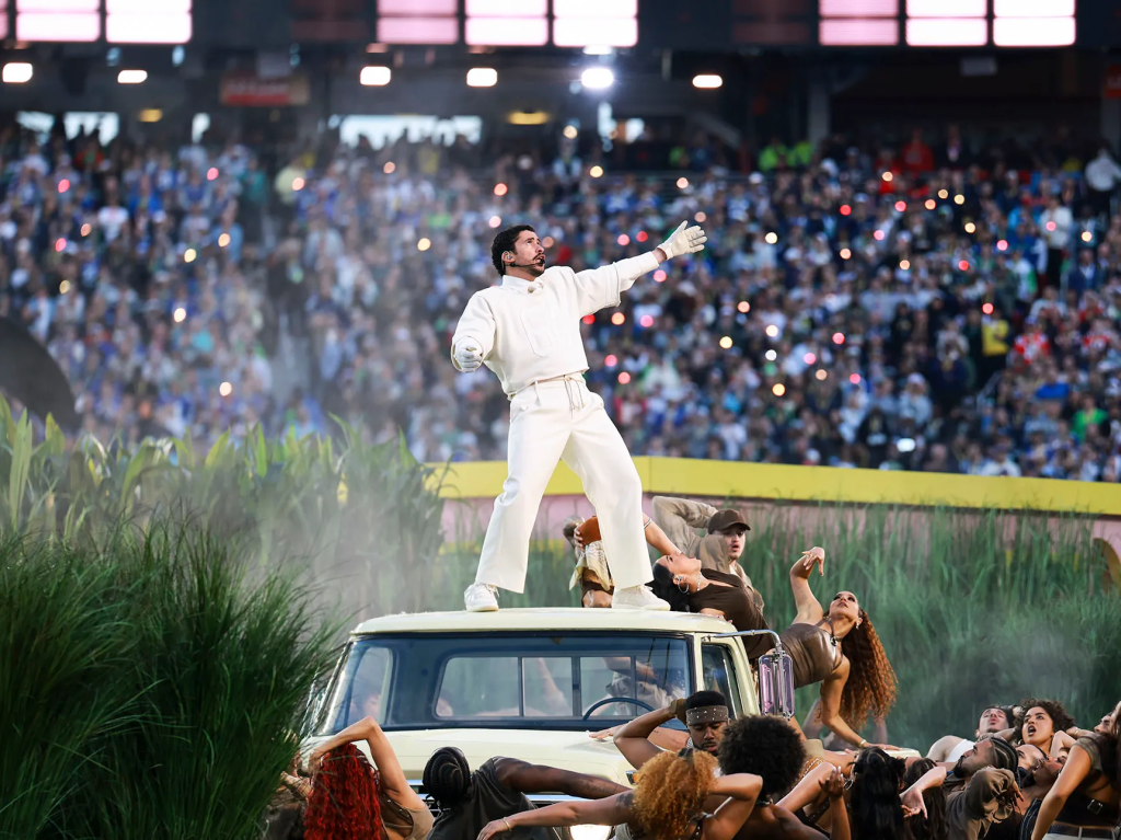 A performer in a white outfit stands on top of a vintage car, surrounded by dancers in a lively outdoor concert, with a crowd of fans and bright lights in the background.